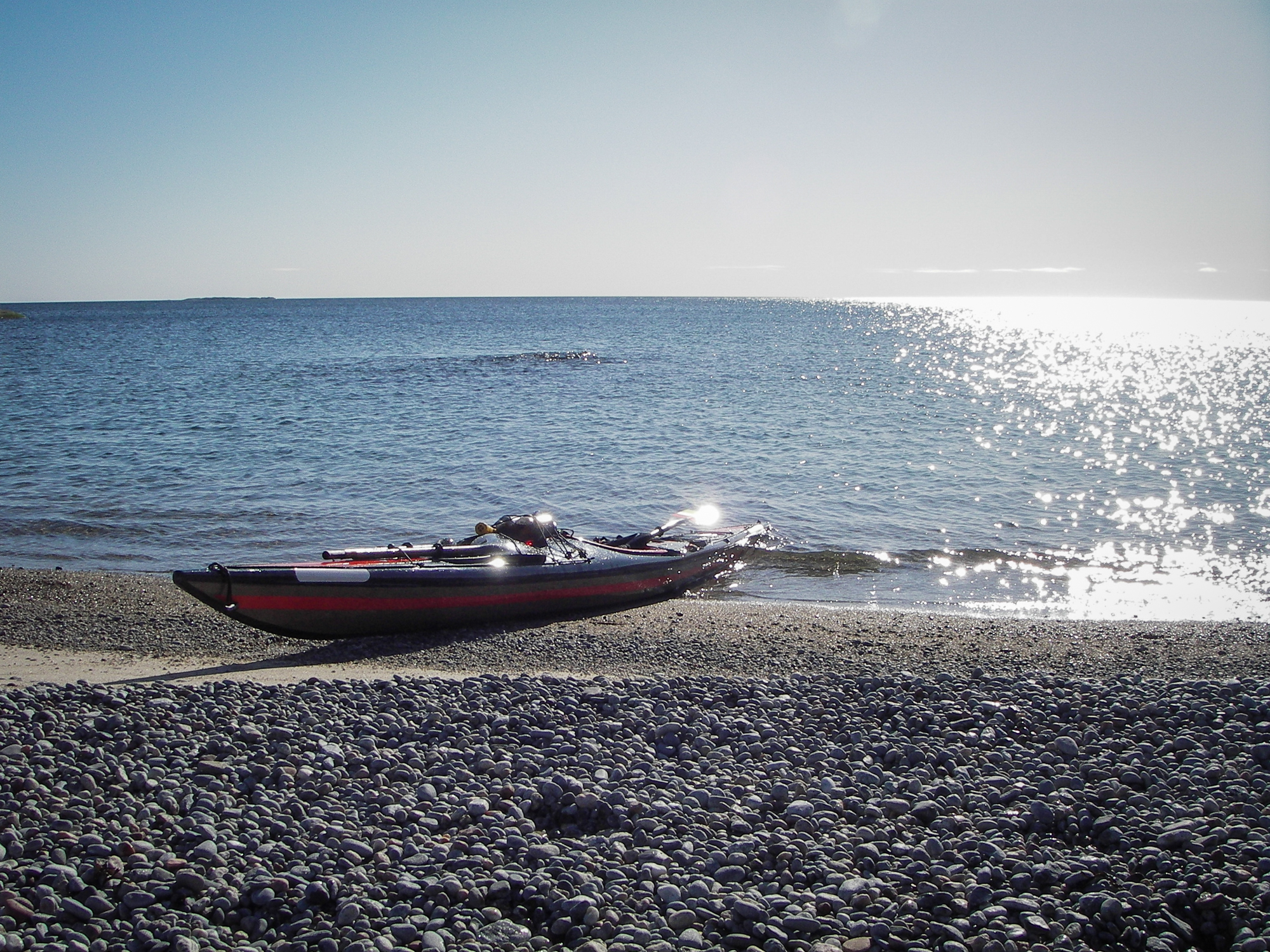 Kajak i strandkanten på stenstrand. Blå himmel och glittrig horisont i bakgrunden.