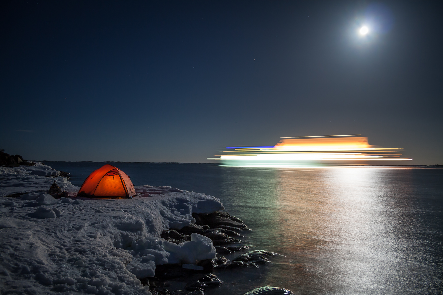 A sea kayakers tent on the ice by the sea in the moonlight with a cruise ship passing by